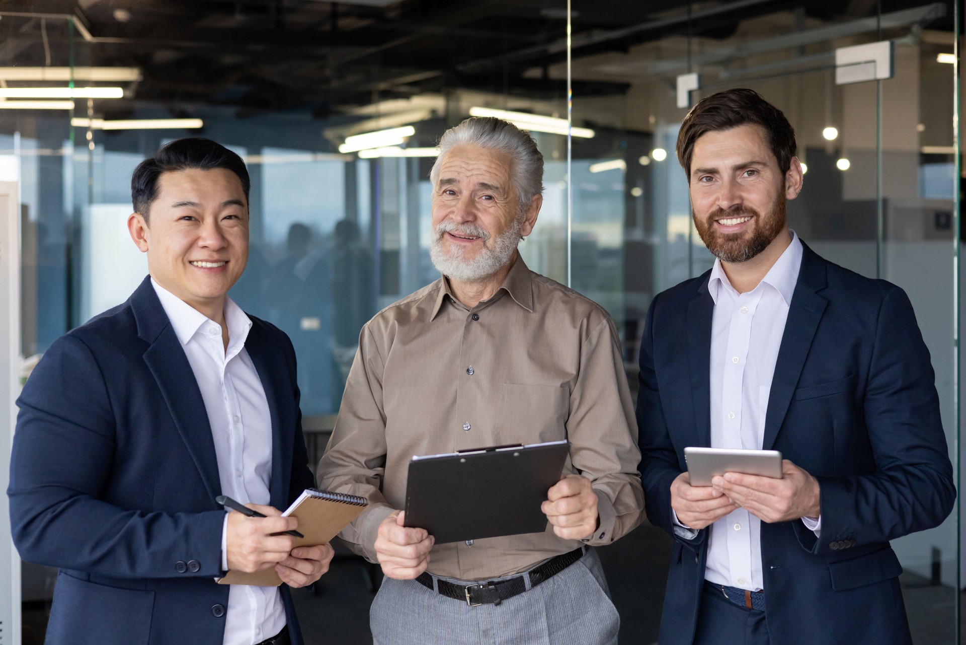 Portrait of three businessmen of different ages and nationalities. They are standing in the office, holding tablets and documents in their hands, confidently and smilingly looking at the camera